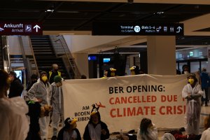 Eine Gruppe maskierter Individuals mit einer Tafel, auf der "Ber Opening Due to Climate Crisis Cancelled" steht, steht vor verstreuten Taschen und Gegenständen, mit Rolltreppen, Texttafeln, Lampen und Säulen im Hintergrund.