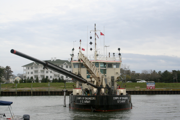 Ein Schiff auf dem Wasser mit Bäumen, Fahrzeugen, Gebäuden und Pfählen im Vordergrund und einem Himmel im Hintergrund, mit einem Boot in der linken unteren Ecke.