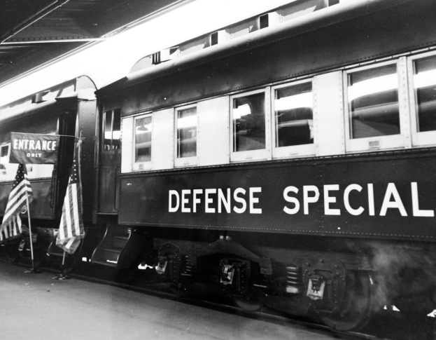 Ein Schwarz-Weiß-Foto eines Zuges mit der Aufschrift "Verteidigungsspezial" auf den Eisenbahnschienen geparkt, mit zwei Fahnen auf der linken Seite und einer Decke darüber.