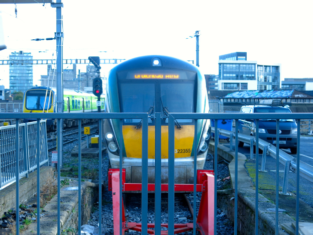Zwei Züge befinden sich auf dem Gleis mit einem Zaun im Vordergrund, einigen Pfählen auf dem Land, einschließlich einem mit einer Signalleuchte, einem Auto auf der Straße rechts, Gebäuden im Hintergrund und dem Himmel darüber.