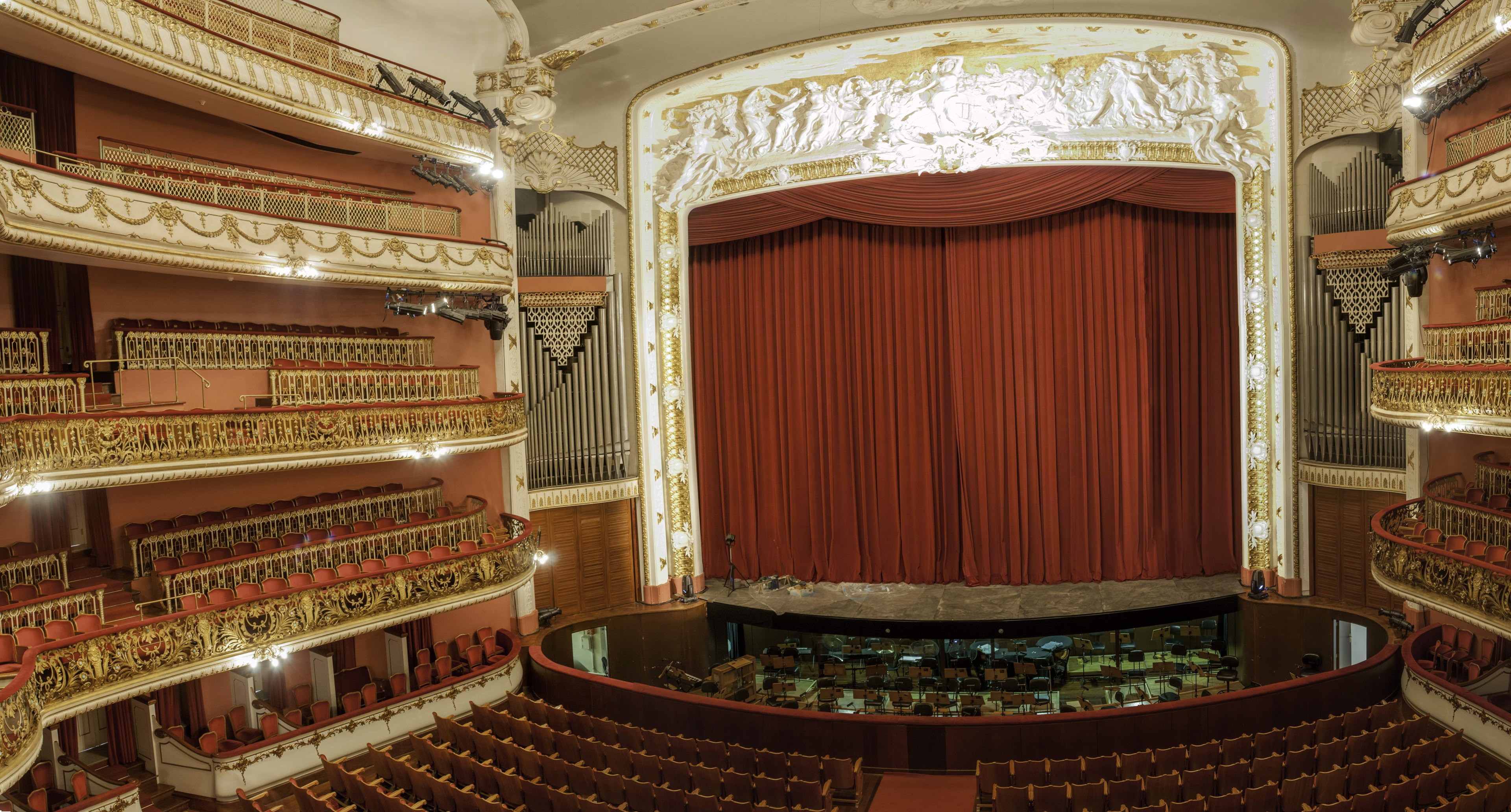 Großer Auditoriumsinnenraum mit roter Gardine, Sitzreihen, Geländern, Beleuchtungskörpern, Wandskulpturen und Bühnenobjekten, die eine Konzertsäleinrichtung suggerieren.