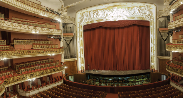 Großer Auditoriumsinnenraum mit roter Gardine, Sitzreihen, Geländern, Beleuchtungskörpern, Wandskulpturen und Bühnenobjekten, die eine Konzertsäleinrichtung suggerieren.