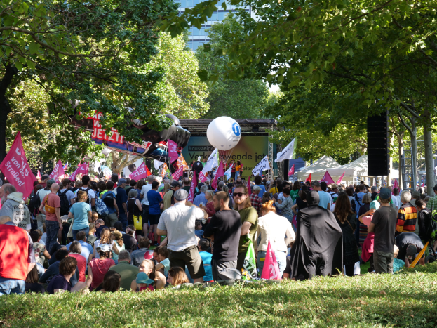 Eine große Menschenmenge sitzt auf einem grünen Feld, einige tragen Kappen und Schutzbrillen, einige halten Fahnen, mit Bäumen, Zelten, Bannern, einem Ballon und einem Gebäude mit Fenstern im Hintergrund.
