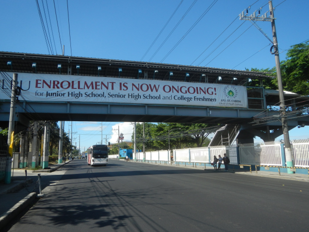 Ein Bus, der unter einer Brücke mit einem "Jetzt Anmelden"-Schild fährt, Fußgänger auf dem Gehweg darunter, Strommasten mit Drähten, Bäume und ein klarer blauer Himmel im Hintergrund.