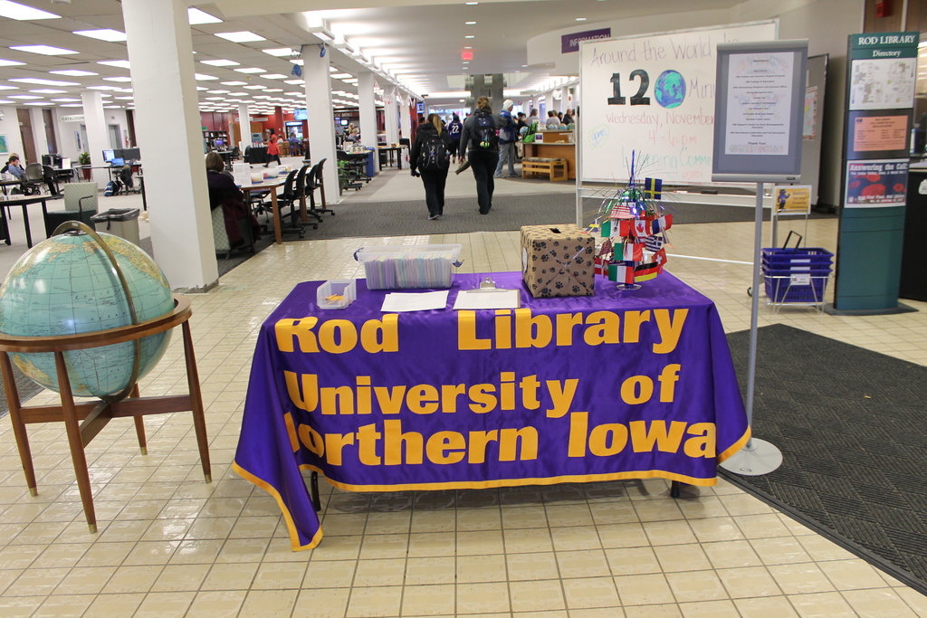 Eine Messestellung mit einem Tisch, auf dem verschiedene Landesflaggen, eine Box, Papiere und ein Banner sowie links daneben eine Weltkugel, rechts daneben eine Anschlagtafel mit einigen Menschen und links daneben viele Tische und Stühle zu sehen sind.