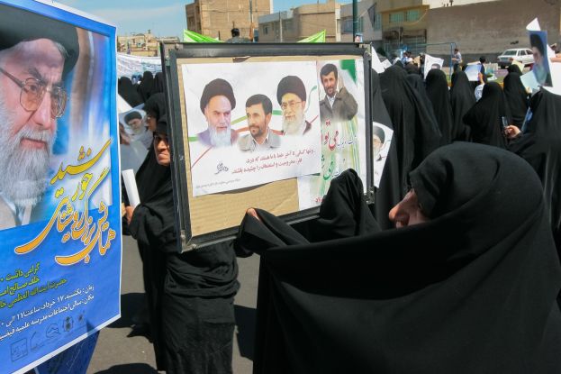 Eine Gruppe von Frauen in schwarzen Abayas hält Plakate mit Fotos von Ayatollah Ali Khamenei vor einem Hintergrund aus Gebäuden, Pfählen, Fahrzeugen und einem klaren blauen Himmel.