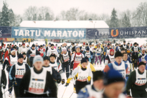 Eine Gruppe von Menschen, die auf Skiern in einem Rennen laufen, tragen Mützen und halten Stöcke, mit Bannern, Bäumen und einem klaren blauen Himmel im Hintergrund und Text am unteren Rand des Bildes.