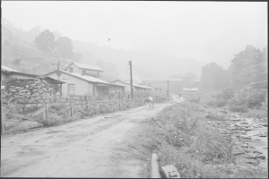 Ein Schwarz-Weiß-Foto einer Schotterstraße mit einem Pferd darauf, umgeben von Pflanzen, Bäumen, Häusern, Pfählen, Drähten und einem Zaun, mit Hügeln und einem klaren Himmel im Hintergrund und Text am unteren Rand des Bildes.