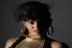 Eine Frau in einem goldenen Kleid und großen Ohrringen steht selbstbewusst vor einer Wand, ihre Haare sind zu einem sleeken Bob gestylt und sie schaut direkt in die Kamera mit einem starken und entschlossenen Ausdruck.