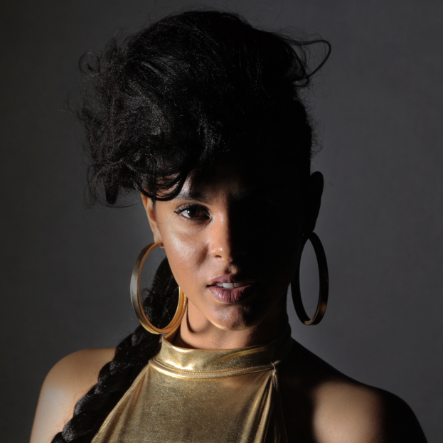 Eine Frau in einem goldenen Kleid und großen Ohrringen steht selbstbewusst vor einer Wand, ihre Haare sind zu einem sleeken Bob gestylt und sie schaut direkt in die Kamera mit einem starken und entschlossenen Ausdruck.