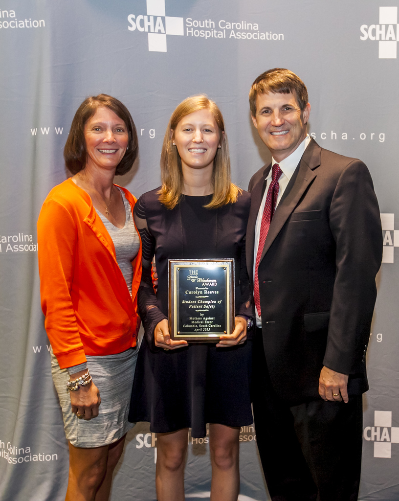 Drei Personen, zwei Frauen und ein Mann, stehen zusammen. Der Mann trägt einen schwarzen Anzug, eine Frau hat ein orange-schwarzes Kleid an und die andere Frau ein schwarzes Kleid und hält eine Trophäe in der Hand. Sie stehen vor einem grauen Hintergrund.
