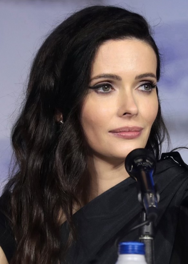 Krysten Ritter auf der San Diego Comic Con, tragend ein schwarzes Kleid, vor einem Mikrofon mit einer Flasche unten, mit einem unscharfen, traumhaften Hintergrund.