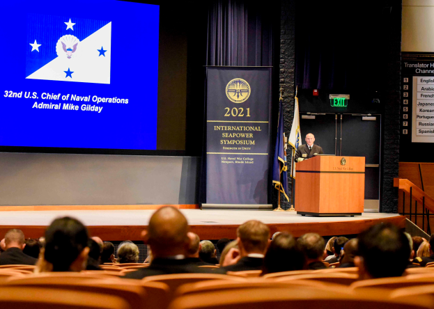 Admiral Mike Gilday steht an einem Podium mit einem Mikrofon und einer Tafel, spricht zu einer sitzenden Menge auf der 2021 International Seapower Symposium, mit Flaggen, einem Bildschirm mit Text und einer Treppe mit Geländern im Hintergrund.