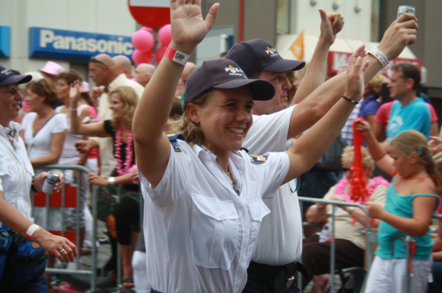 Eine Gruppe von Menschen steht und sitzt vor einer Menge bei einem Umzug, einige tragen Mützen und halten Dosen, mit Geländern, Luftballons und einem Gebäude mit Namensschildern im Hintergrund.