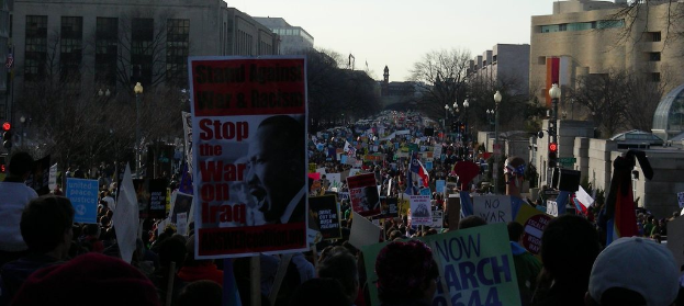 Menschen mit Plakaten auf einer Straße, mit Gebäuden, Bäumen, Laternenpfählen und dem Himmel im Hintergrund.