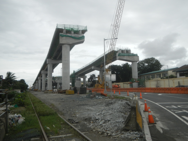 Baustelle mit einer Brücke im Hintergrund, Straße durch Verkehrskegel markiert, Eisenbahnschiene auf der linken Seite, verstreute Steine und Gras, Bäume und Gebäude auf beiden Seiten der Straße und ein bewölkter Himmel.