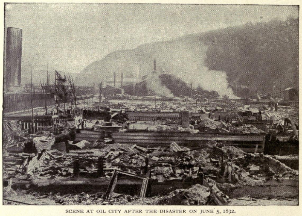 Schwarzes und weißes Foto einer Stadt nach einer Katastrophe am 5. Juni 1892, das Gebäude, Pfosten, Rauch und einen Hügel im Hintergrund zeigt, mit Text unten.