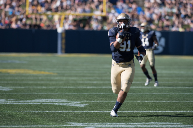 Ein Fußballspieler mit Helm rennt mit dem Ball auf einem Feld, umgeben von einer unscharfen Menge.