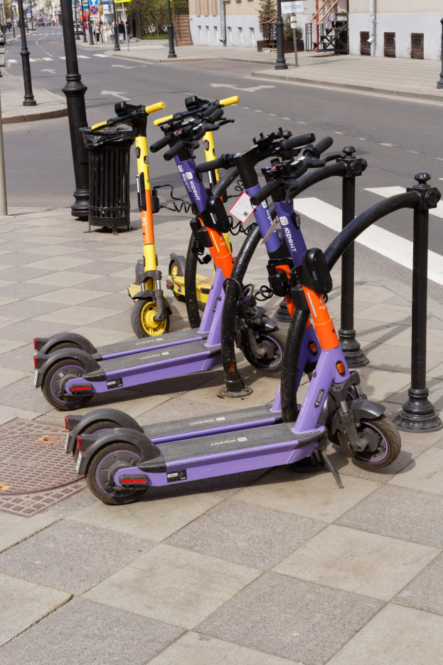 Eine Reihe von Elektro-Scootern, die entlang einer Straße geparkt sind, mit einem Mülleimer im Vordergrund, Gebäude, Bäume und Fahrzeuge im Hintergrund.