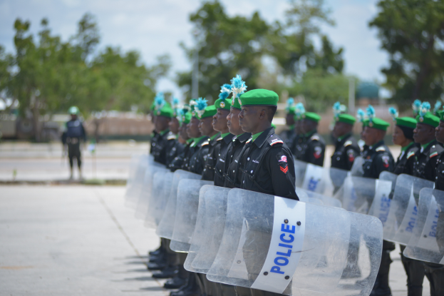 Eine Reihe von Polizisten in schwarzen Uniformen und grünen Mützen hält durchsichtige Schilde, mit Bäumen, Gebäuden und einem klaren blauen Himmel im Hintergrund.