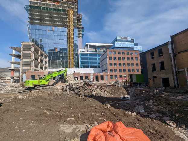 Großbaustelle mit teilweise errichteter Struktur, Kränen, verstreuten Steinen und Schutt unter einem bewölkten Himmel, mit einem Fertigstellungstermin am 20. April 2020.