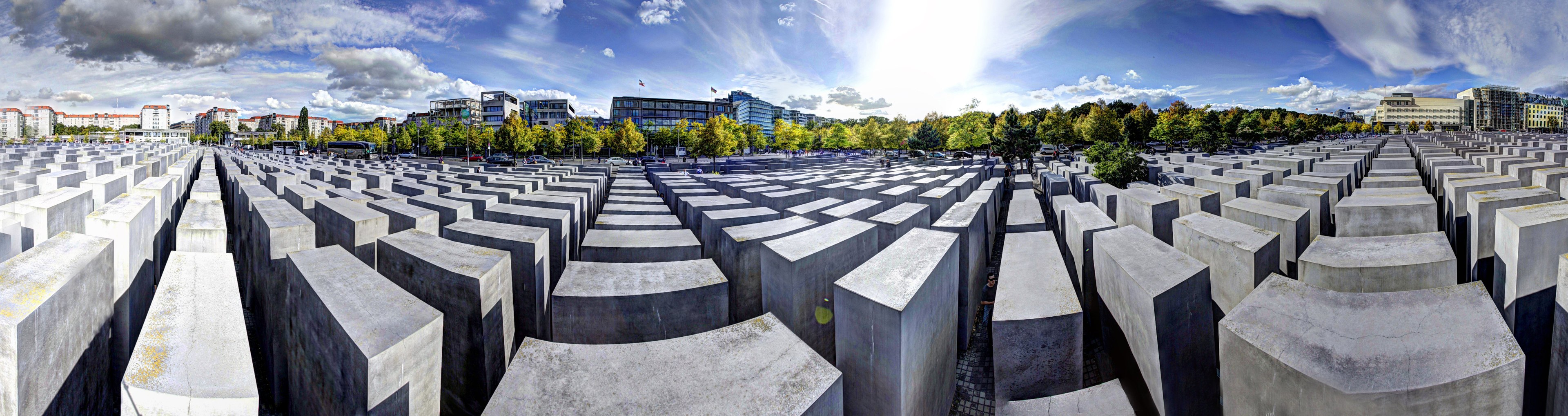 Großes Beton-Mahnmal für die ermordeten Juden Europas in Berlin, umgeben von Bäumen und Gebäuden, mit Fahrzeugen auf der Straße und einem bewölkten Himmel.