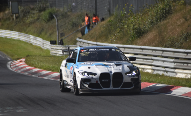 Ein BMW M8 GT3 Rennwagen auf einer Rennstrecke, umgeben von einem Zaun, Gras, Pflanzen und einer Gruppe von Menschen, mit sichtbarem Text auf dem Auto und einem unscharfen Hintergrund.