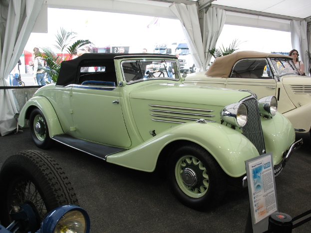 Mehrere Oldtimer sind auf einem Boden mit Vorhängen und Pflanzen im Hintergrund geparkt, während weiter hinten auf der Straße einige Menschen und Fahrzeuge zu sehen sind.