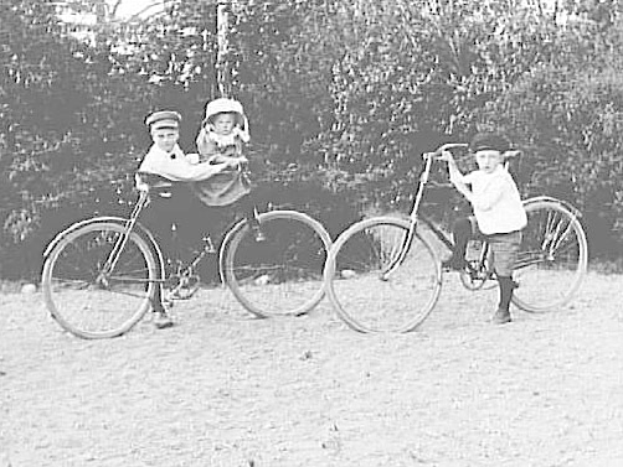 Zwei Kinder fahren Fahrräder in einem Park, eines hält ein Baby, mit Bäumen und einem Pfahl im Hintergrund, in Schwarz-Weiß.
