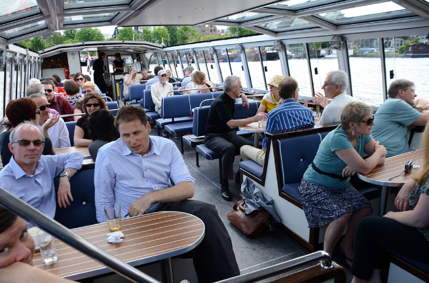 Ein Boot-Innere mit Sitzreihen, auf denen sich Menschen befinden, Tischen mit Gläsern und Schüsseln davor, einer Tasche auf dem Boden, Wasser draußen sichtbar, Gebäuden im oberen rechten Hintergrund und kleinen Pflanzen über dem Boot.