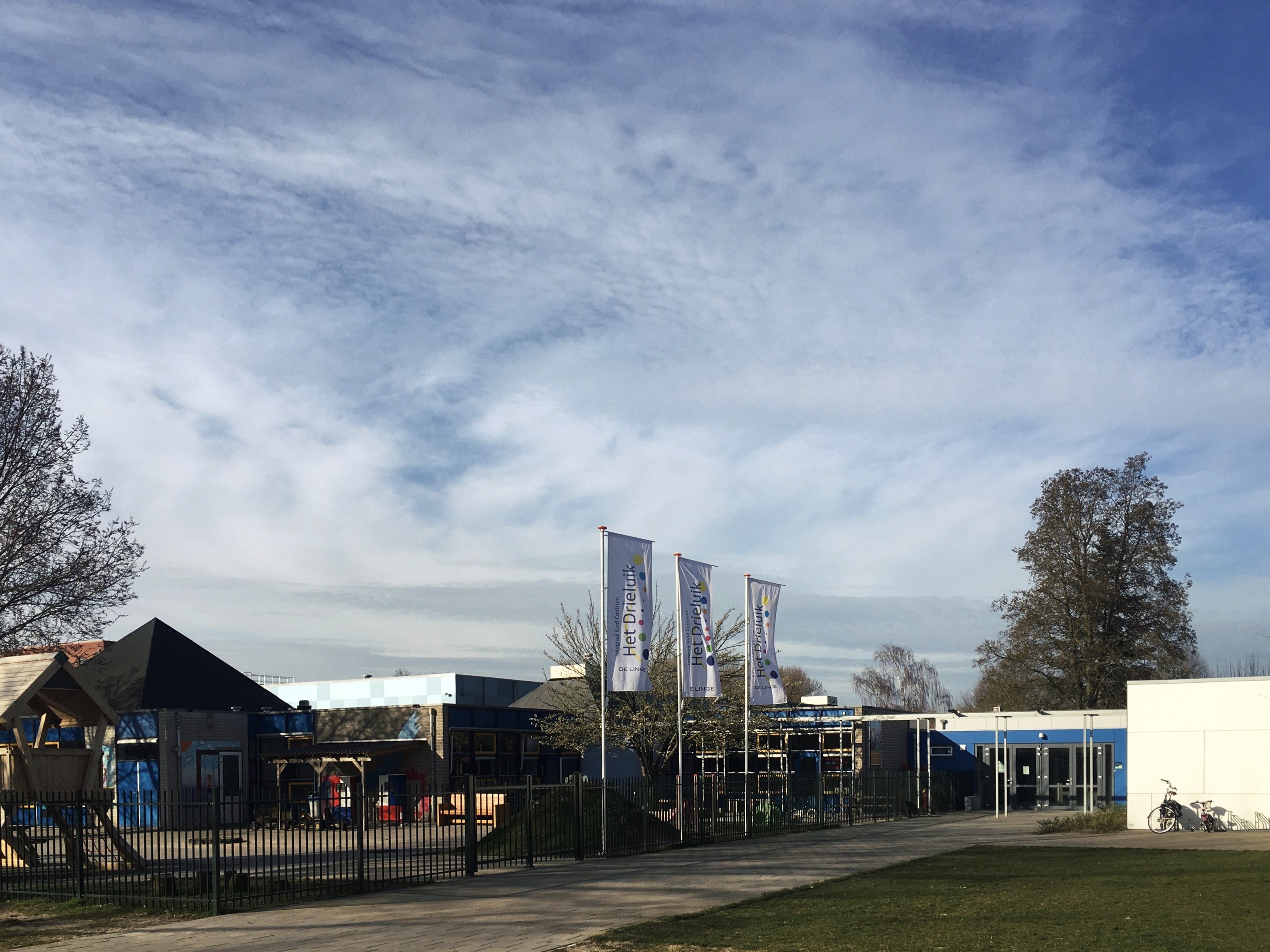 Großes Schulgebäude mit Metallzaun, Rasenfläche, Weg, Bäumen, Bannern, einem geparkten Fahrrad, nahen Häusern und einem bewölkten Himmel, als neuer Spielplatz für Kinder bezeichnet.