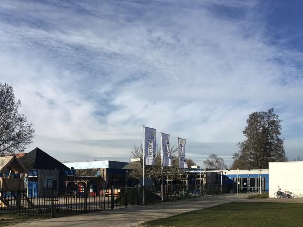 Großes Schulgebäude mit Metallzaun, Rasenfläche, Weg, Bäumen, Bannern, einem geparkten Fahrrad, nahen Häusern und einem bewölkten Himmel, als neuer Spielplatz für Kinder bezeichnet.