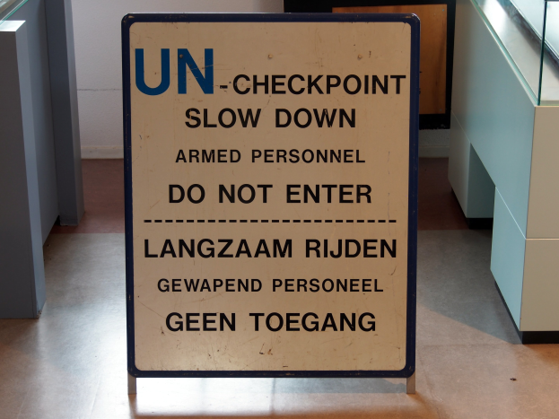 Schild auf dem Boden mit der Aufschrift "UN-Checkpoint Verlangsame bewaffnete Personen - Betreten verboten" mit einem Glastisch und Gegenständen rechts daneben und einer Wand mit einem Gegenstand im Hintergrund.