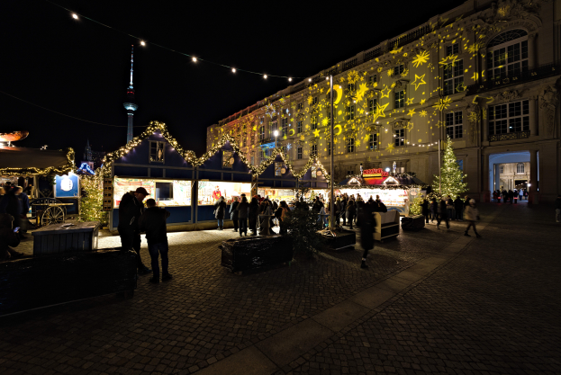 Ein lebendiger Weihnachtsmarkt in Berlin, Deutschland, mit Menschen um beleuchtete Stände, festliche Dekorationen und dunklem Himmel im Hintergrund.
