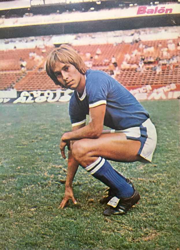 Ein Mann kniet auf einem Fußballfeld, umgeben von sitzenden Menschen mit Stühlen und Tafeln mit Text, trägt Schuhe.