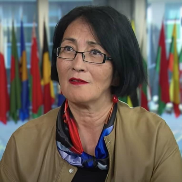 Eine Frau mit Brille und Schal sitzt vor einer Wand mit Flaggen und spricht während eines Interviews über Frauenrechte.