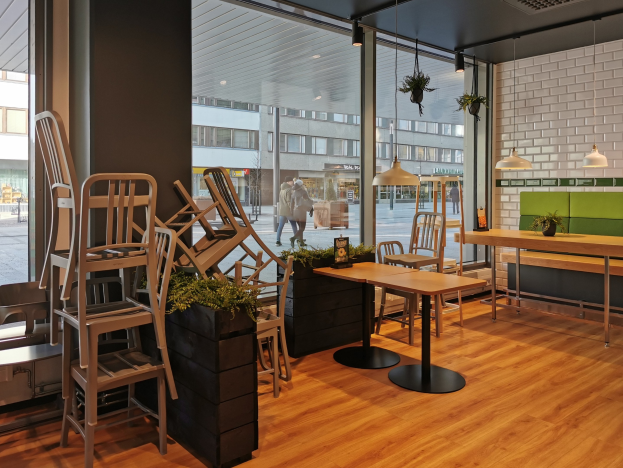 Ein Restaurant-Innenraum mit hölzernen Tischen und Stühlen in der Nähe eines großen Fensters, das auf eine Berliner Straße mit Fußgängern, Gebäuden und städtischen Elementen blickt.