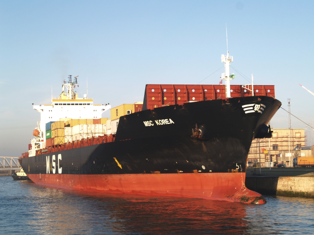 Ein großes Container-Schiff liegt neben einem Dock mit einer Brücke, Gebäuden, Pfählen und einem Kran auf der rechten Seite, unter dem Himmel.