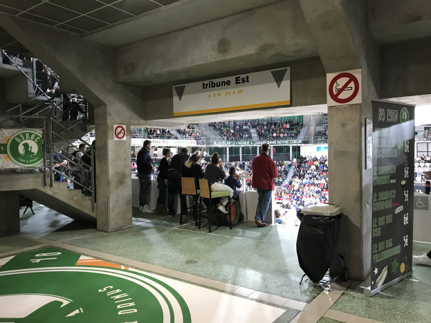 Eine Gruppe von Menschen steht vor einem sitzenden Publikum in einem Stadion, mit Schildern, die "Boston Celtics vs New York Jets" lesen, einem Müllcontainer, einer Tafel mit Text, einer Treppe mit Geländern und Deckenleuchten.