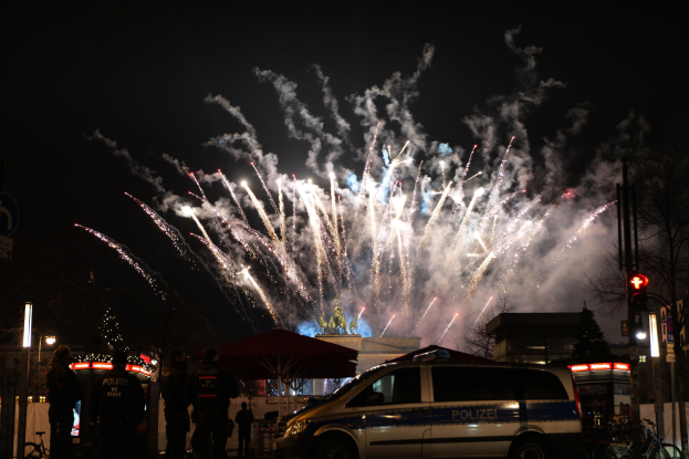 Polizeiauto vor einer Menschenmenge während einer Silvesterfeier in Berlin geparkt, mit farbenfrohen Feuerwerken, die den Himmel erleuchten und die umliegenden städtischen Elemente.