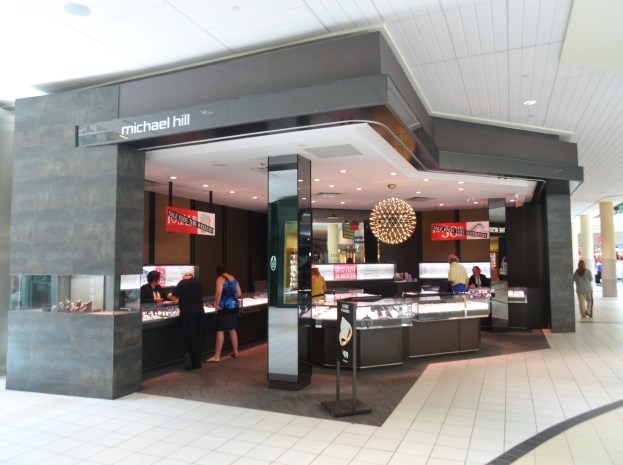 Ein Michael Hill Schmuckgeschäft in einem Einkaufszentrum, mit ein paar Menschen davor, beleuchtet von Deckenlampen.