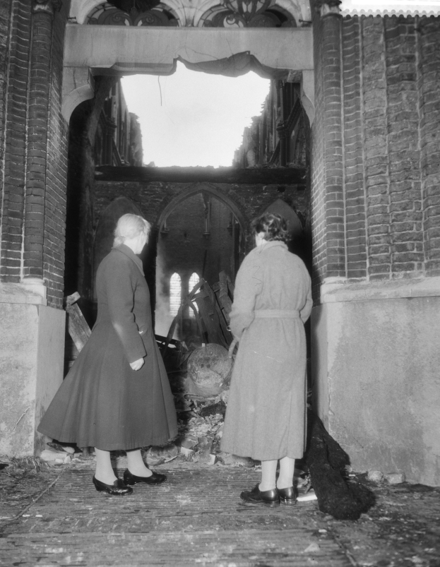 Zwei Frauen stehen in der Tür eines feuerbeschädigten Gebäudes, umgeben von verkohlten Überresten, dargestellt in Schwarz-Weiß.