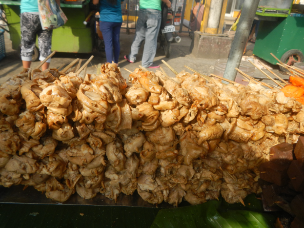 Ein Straßenfood-Stand mit Hühnerkebab auf Bananenblatt, Menschen, ein Fahrzeug, ein Pfahl und ein Tisch mit verschiedenen Gegenständen auf der linken Seite.