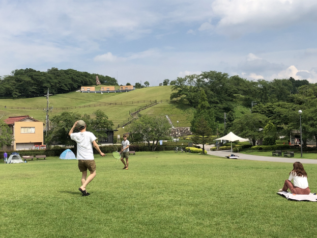 Gruppe von Menschen, die Badminton in einem Park spielt, mit einem Mann und einer Frau auf einer Decke auf dem Rasen sitzend, die Rackets halten, Zelte und Gebäude im Hintergrund.