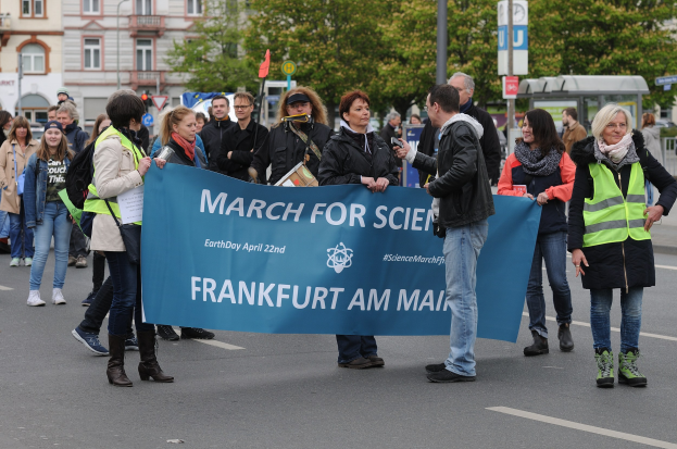 Eine bunte Gruppe von Menschen marschiert die Straße entlang, trägt ein Banner mit der Aufschrift "March for Science Frankfurt am Main" mit Bäumen, Pfählen, Schildern, Gebäuden und einem klaren blauen Himmel im Hintergrund.
