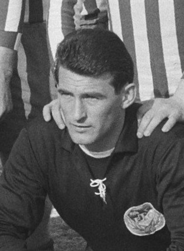 Schwarzes und weißes Foto eines Mannes, der auf einem grasbewachsenen Füüballfeld sitzt und ein T-Shirt und eine Kette trägt, mit zwei stehenden Figuren hinter ihm.