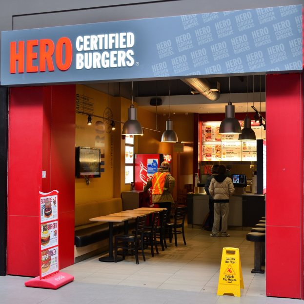 Ein Hero Certified Burgers Restaurant im Innern von London mit Menschen, Tischen, Stühlen, einer Speisekarte, einem wandmontierten Fernseher und Deckenleuchten.