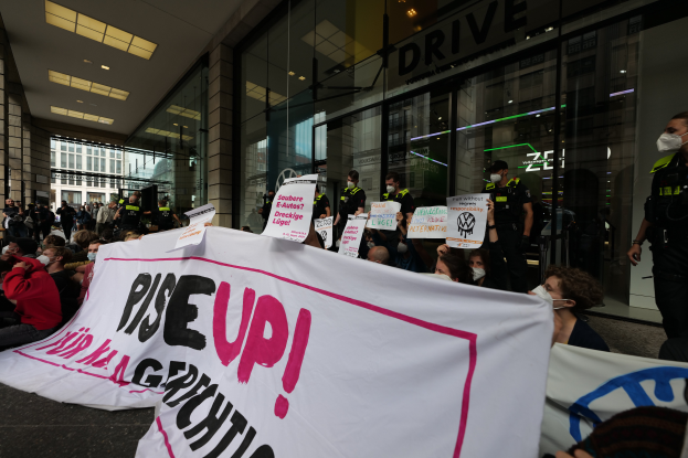 Eine Gruppe von Menschen h"lt Schilder und Plakate mit der Aufschrift "Steh Auf" und protestiert vor einem Geb"ude mit Glasw"anden und Deckenleuchten, einige tragen M"asken.