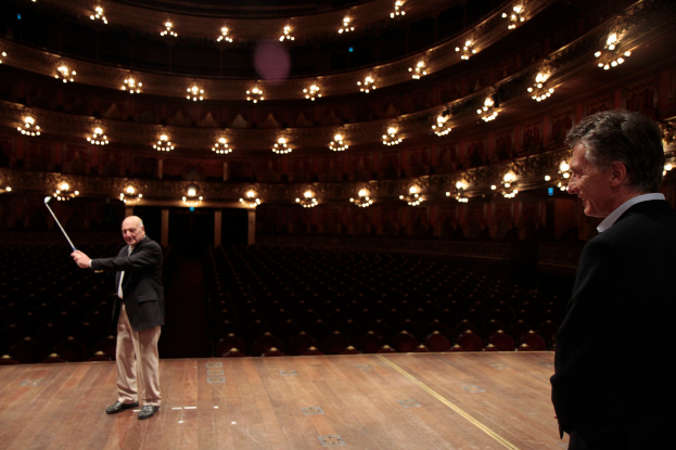 Ein älterer Mann im Anzug hält einen Golfschläger auf der Bühne, neben ihm steht ein weiterer älterer Mann unter einer Deckenbeleuchtung mit vielen Sitzplätzen darunter.