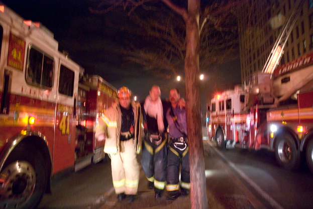 Drei Personen vor einem Feuerwehrauto auf der linken und einem Gebäude auf der rechten Seite mit einem Baum in der Mitte.
